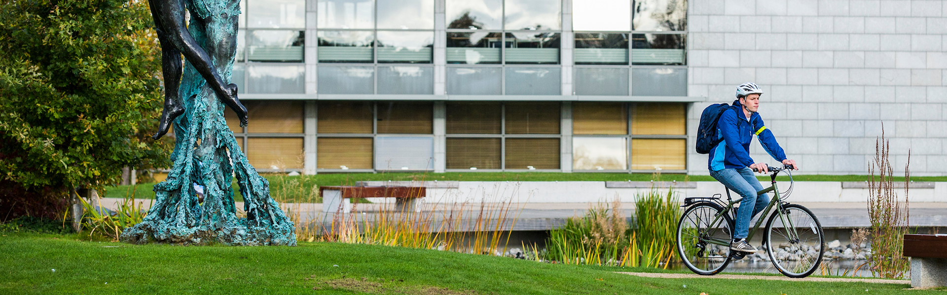 Young man cycles his bicycle past a lake with reads. A grey building with larger windows is in the background.